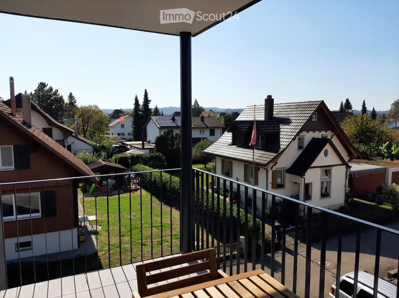 Two-story house with brown and white exterior, balcony with metal railing, wooden table and chair, nearby house with solar panels, greenery and road