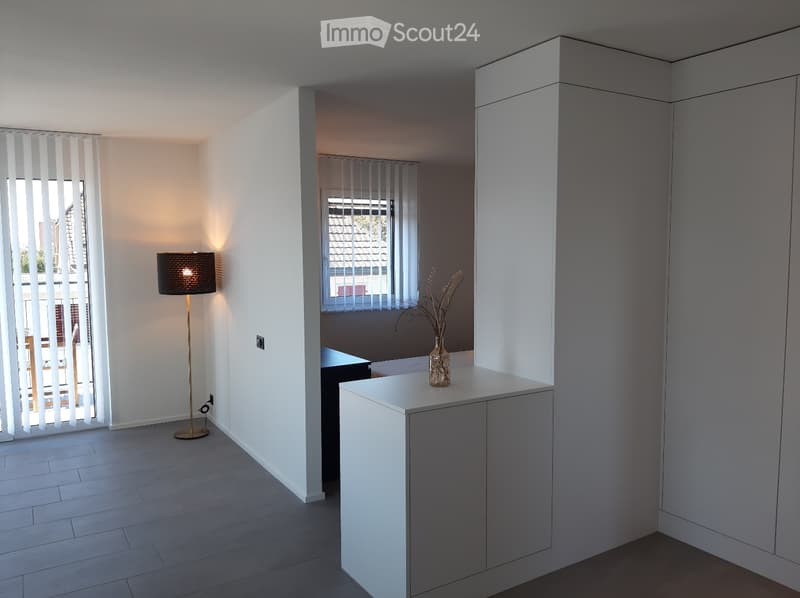 Minimalist room with white walls, tiled floor, a white cabinet with a vase, a lamp, and an open window with blinds. Adjacent room has built-in shelves and a window. Outside view of trees and buildings. A power outlet is on the wall.
