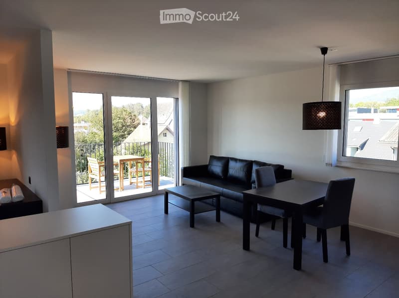 open plan living room with modern black leather sofa, glass doors leading to balcony, dining table, black chairs, pendant lamp, white tiled floor, modern kitchen cabinetry