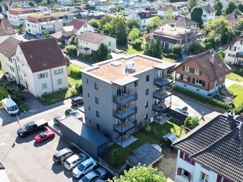Apartment building, multiple cars parked, balconies on sides, surrounding houses, greenery