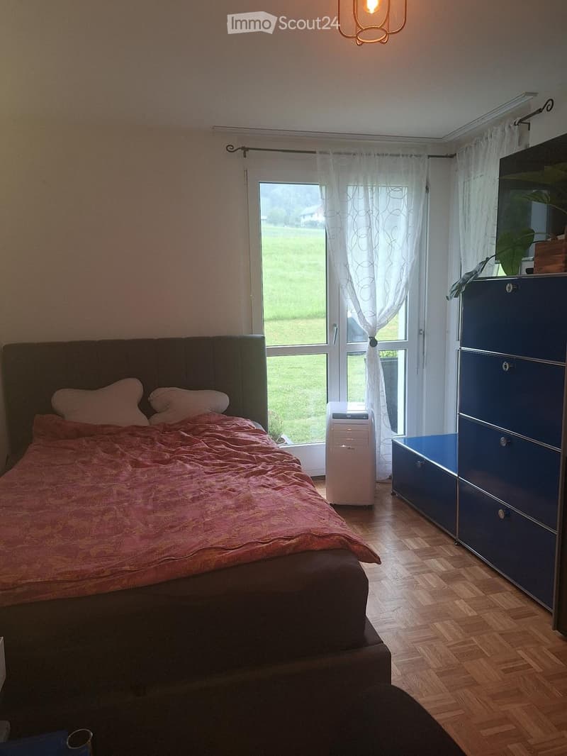 Cozy bedroom with a large bed, headboard, pillows, and a pink blanket. A blue wardrobe with drawers is positioned near a large window with sheer curtains, allowing natural light to fill the room. An air conditioner unit is placed on the floor near the win