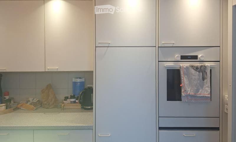 Modern kitchen with white cabinets, a built-in oven, and a refrigerator. The counter has various items including a kettle, bottles, and a bag.