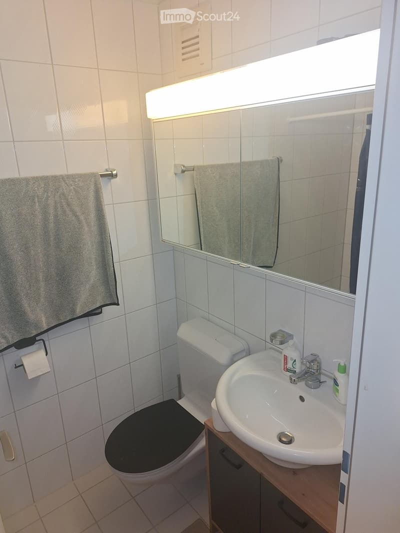 A small bathroom with white tiled walls and floor, featuring a toilet with a black seat cover, a sink with a wooden cabinet, a mirror with built-in lighting, and towels hanging on racks.