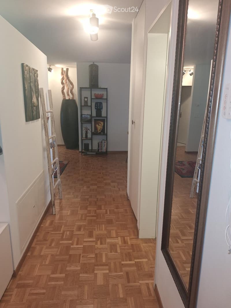 Hallway with wooden herringbone flooring, white walls, and a shelf with decorative items including a vase and a painting. A large mirror is mounted on the right wall.