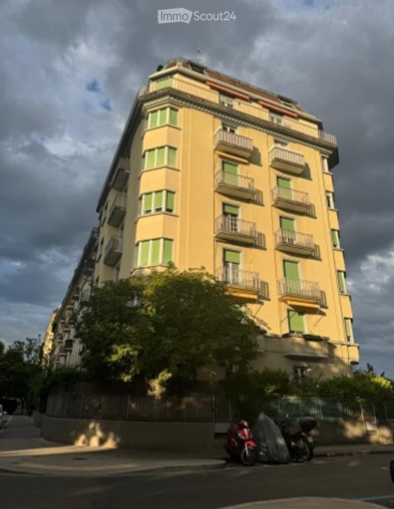 A multi-story building with yellow exterior and green shutters on the windows. The building has multiple balconies and is surrounded by trees and a fence.