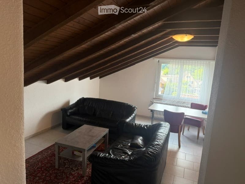 A cozy living room with a slanted wooden ceiling, two black leather sofas, a coffee table, and a dining area with a table and chairs.