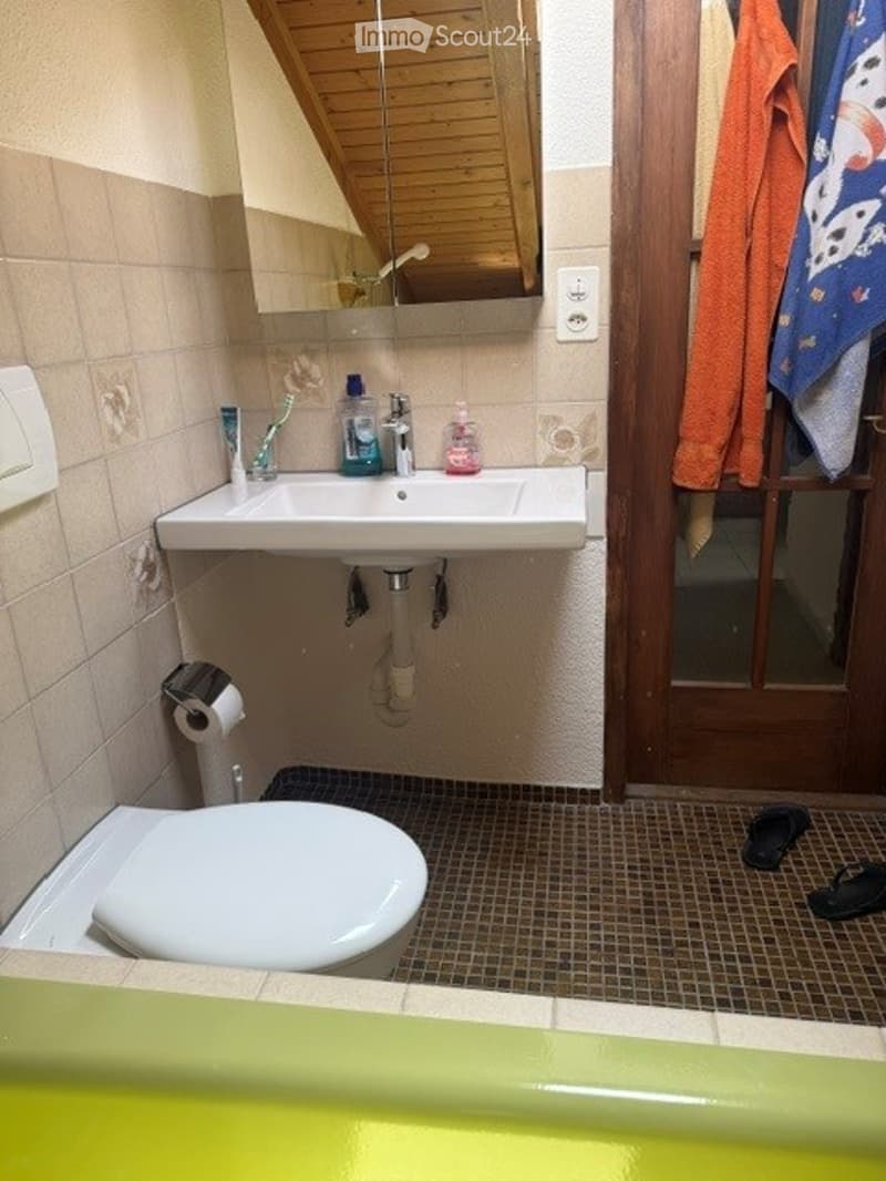 A compact bathroom with a white sink, toilet, and mirror. The sink has a faucet and various toiletries, including a toothbrush and toothpaste. A towel hangs on the door, and a pair of slippers is placed on the floor.