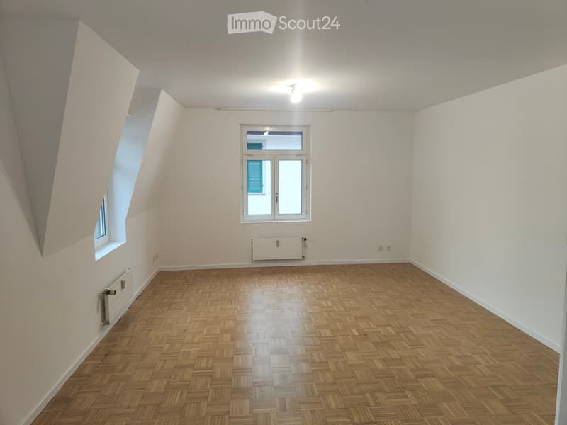 Empty room with wooden herringbone flooring, white walls, and two windows, one with blue shutters visible through the glass.