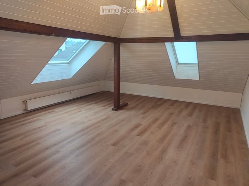 Empty attic space with wooden flooring, white paneled walls, exposed wooden beams, and skylight windows allowing natural light.