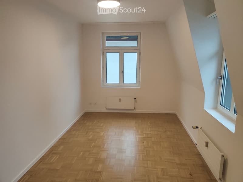 Empty room with wooden herringbone flooring, white walls, and two windows, one with a double door and the other a single window, both with frosted glass panels.