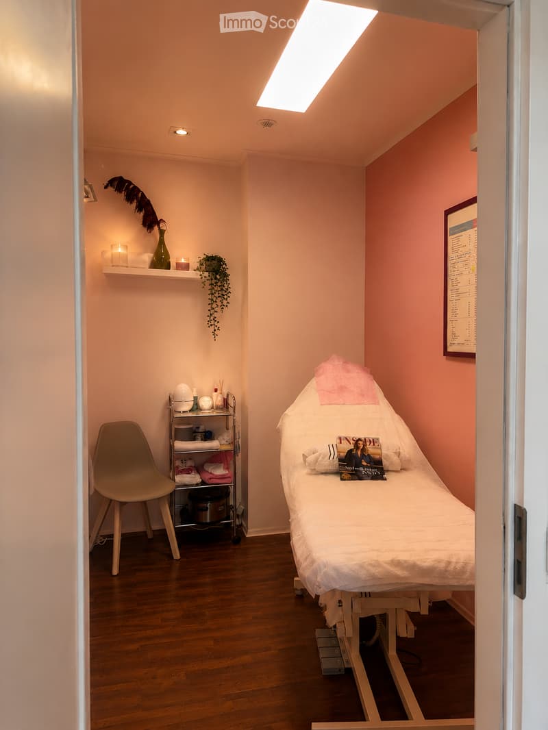 A well-lit, cozy treatment room with peach-colored walls, a wooden floor, a massage bed with white linens, a small shelf with towels and toiletries, a chair, and decorative items including a vase and candles.