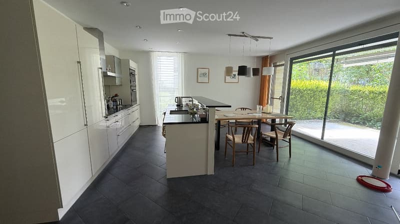 Modern kitchen with white cabinets, dark tiled floor, central island with sink, pendant lights, and a dining area with a table and chairs near large windows.