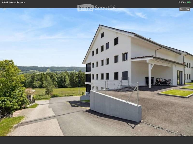 Modern white building, multiple windows, bicycles parked, concrete driveway, chain-link fence, green lawn, trees, mountains in the distance