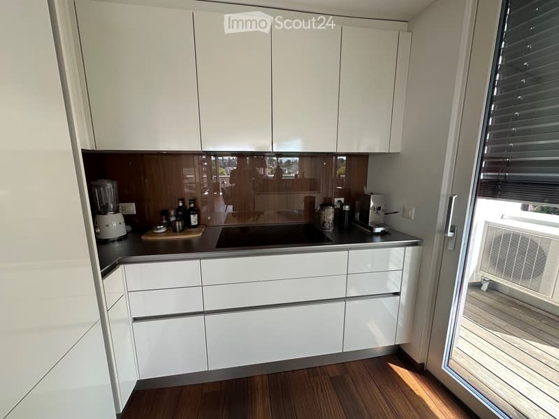 Modern kitchen with white cabinets, dark countertops, and various appliances including a blender and coffee maker. The space features wooden flooring and a large window with blinds.