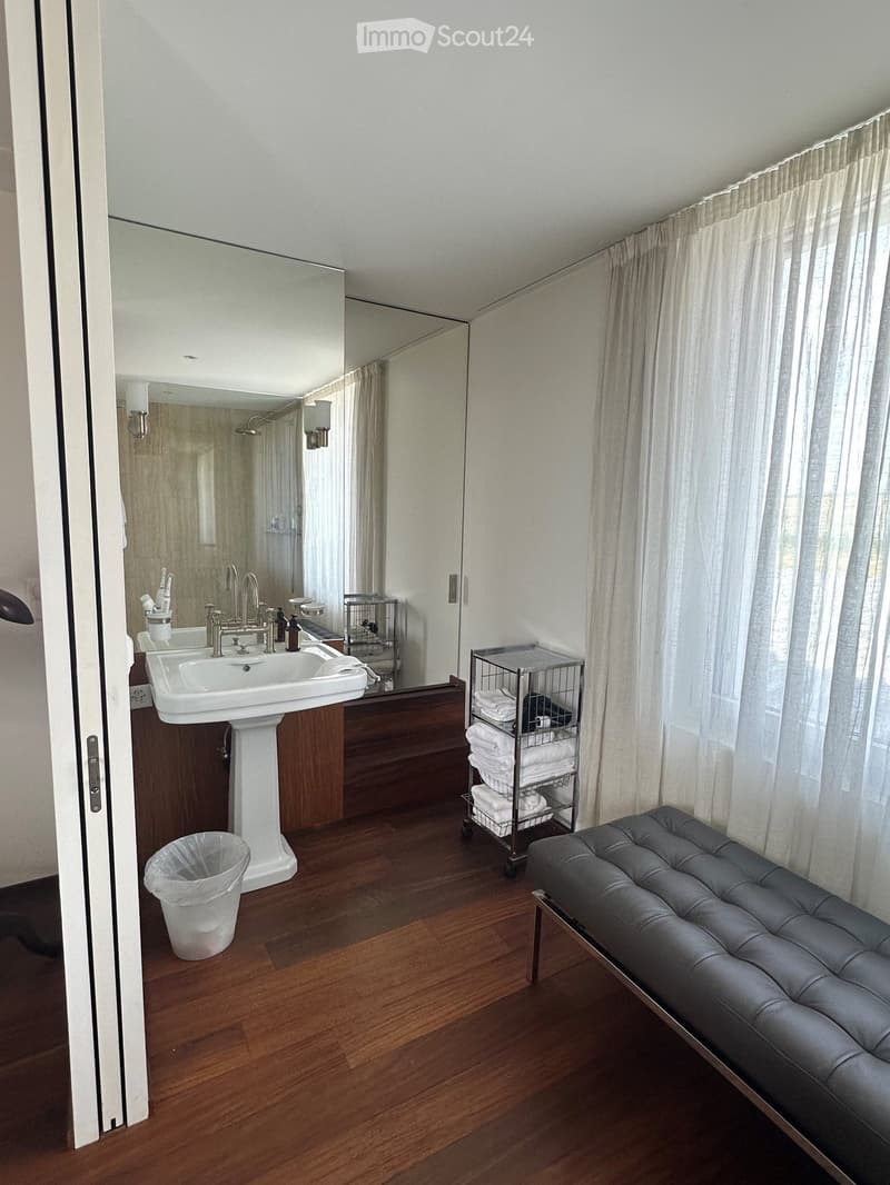 Modern bathroom with wooden flooring, a white sink, a large mirror, a grey leather bench, and a towel cart. The room features large windows with white curtains and a small trash bin near the door.