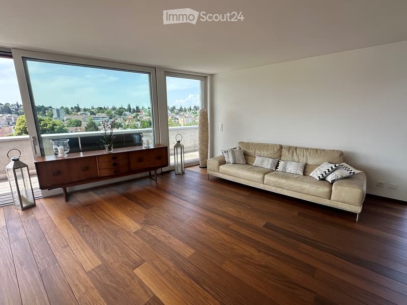 Modern living room with hardwood floors, large windows offering a city view, a beige sofa with patterned pillows, a wooden sideboard, and decorative lanterns.