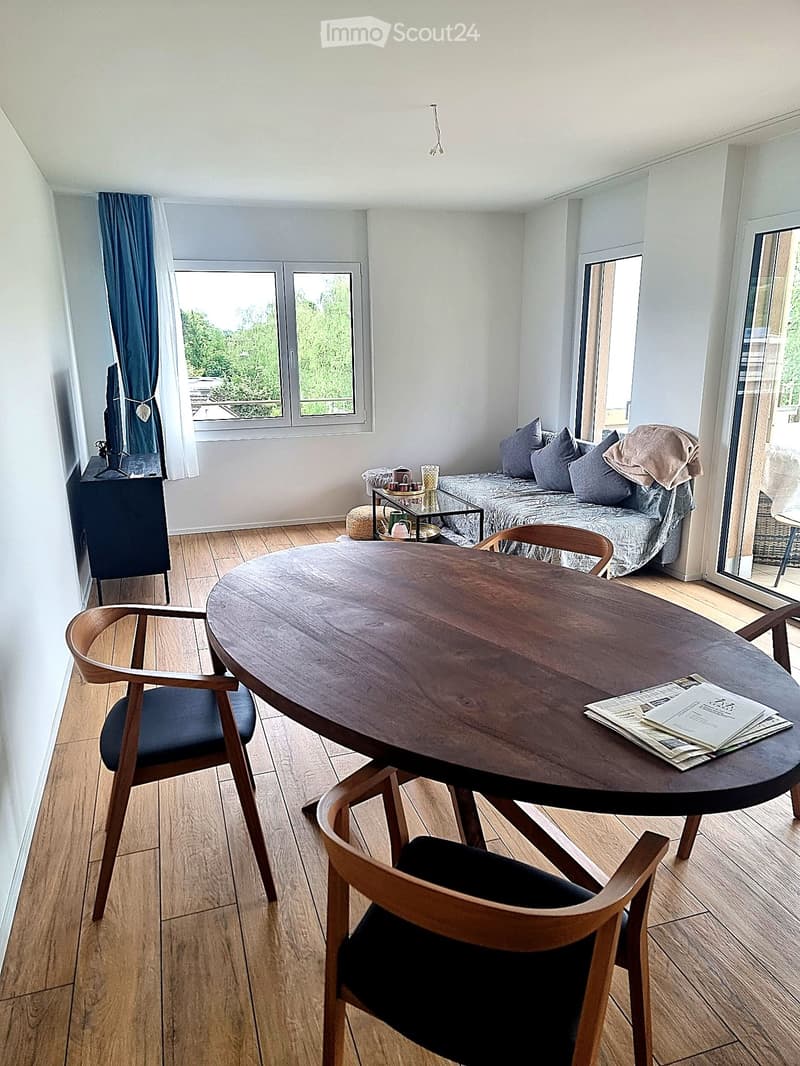 A modern, well-lit room with wooden flooring, a round wooden table with chairs, a sofa with pillows, a television on a stand, and a small side table with cups and a stool.