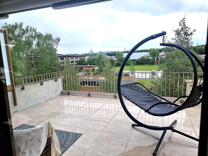 Balcony with tiled floor, metal railing, and a view of greenery and buildings. Features a lounge chair and a small table.