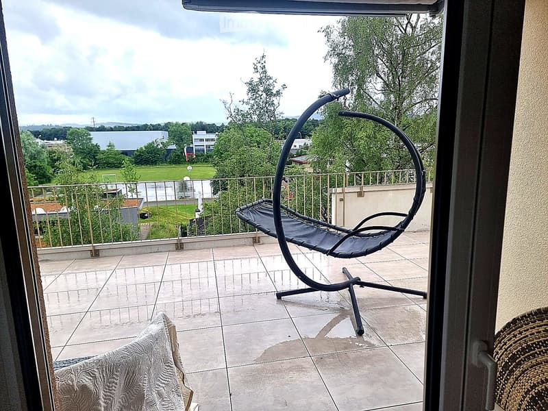 Balcony with tiled floor, metal railing, and a hanging egg chair. Offers a view of a green park and buildings.