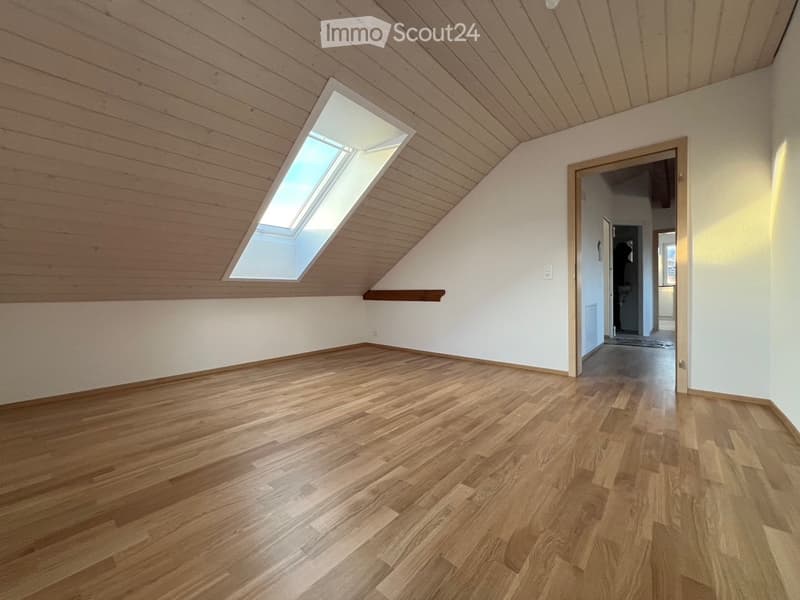 Empty attic room with wooden flooring, white walls, and a skylight window. The room features a slanted wooden ceiling and a small built-in shelf.
