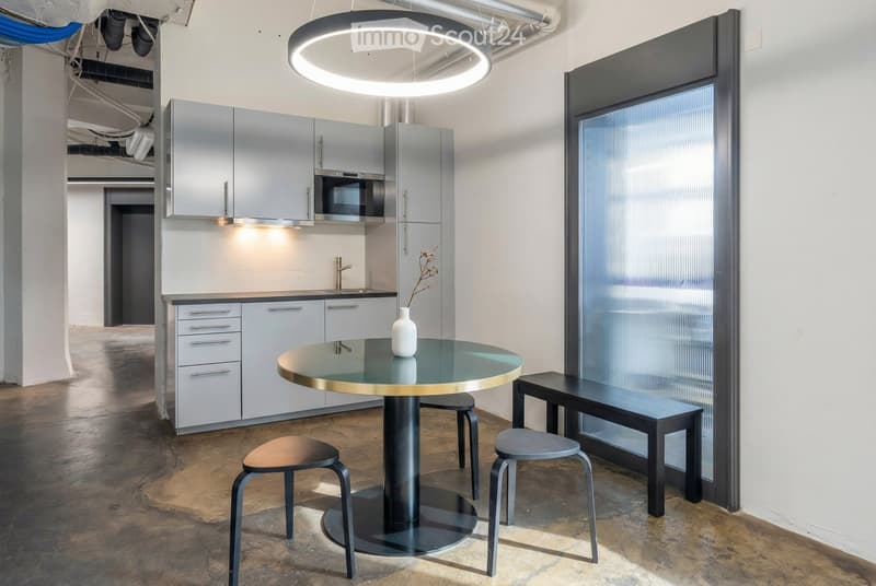 Modern kitchen and dining area with sleek grey cabinets, a round glass table with a gold rim, three stools, a bench, and a vase on the table. The space features exposed pipes and a large circular pendant light.