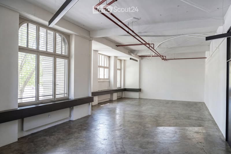 An empty, industrial-style room with concrete floors, white walls, large windows with shutters, and exposed metal pipes on the ceiling.