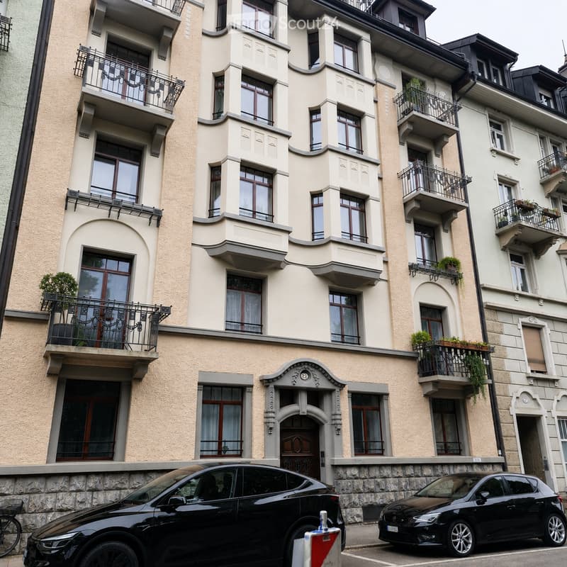 Multi-story residential building with beige facade, multiple balconies, large windows, and decorative stonework. Two black cars are parked in front.