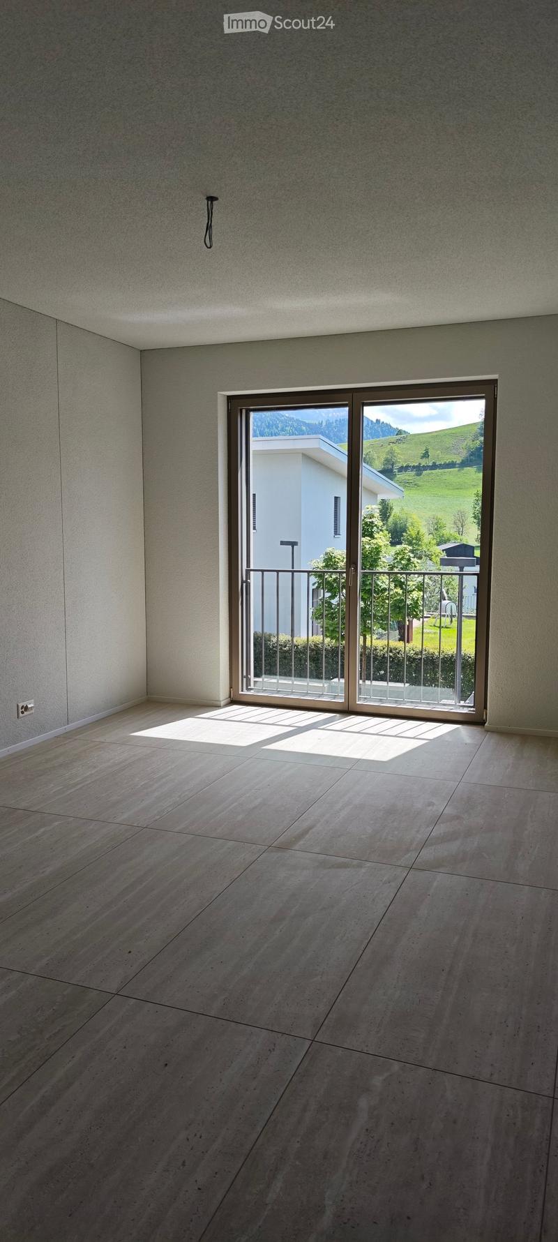 Empty room with light-colored tiled floor and large sliding glass door leading to a balcony with a view of green hills and a neighboring house.