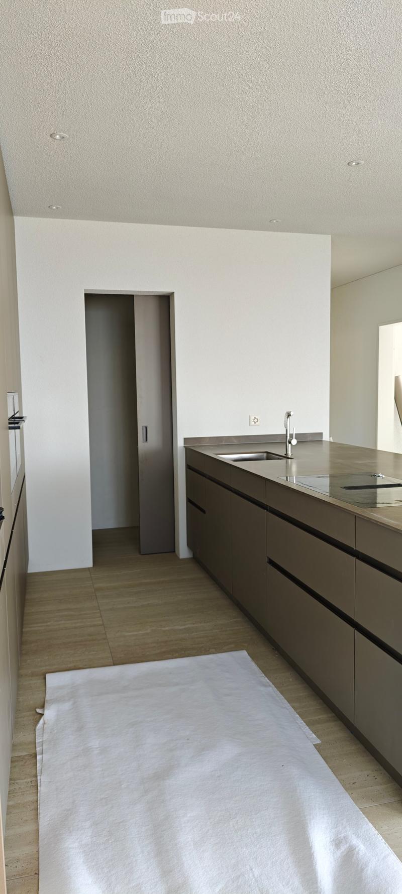 modern kitchen with wooden floor, white walls, stainless steel countertops, double sink, cabinets, and a door leading to another room