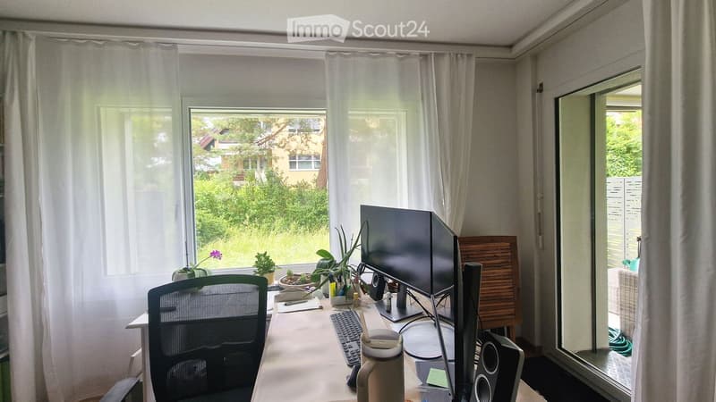 A well-lit home office with a desk, computer monitor, keyboard, chair, and several potted plants. Large windows with sheer curtains offer a view of greenery outside.