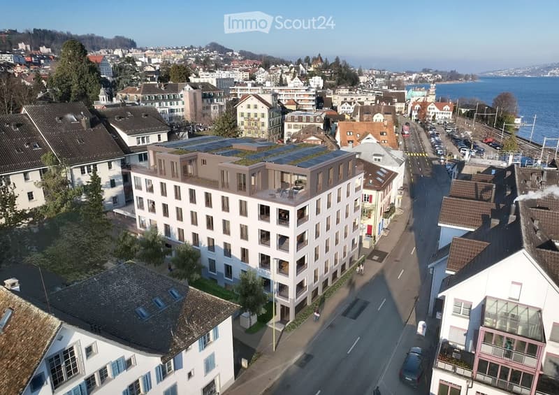 Aerial view of a modern multi-story building with balconies and solar panels on the roof, situated in a residential area near a waterfront with other houses and a road.