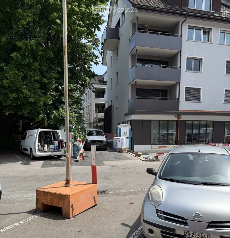 Multi-story residential building with balconies, surrounded by parked cars and construction materials.
