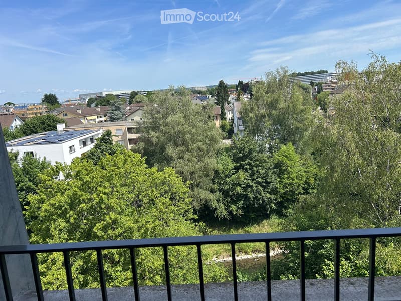 View from a balcony showing a lush green landscape with trees and a variety of buildings in the distance under a clear blue sky.