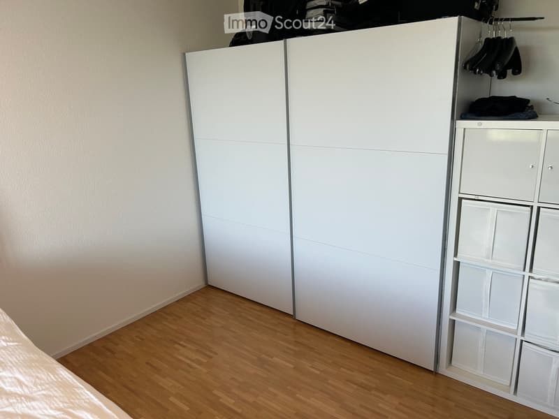 A modern bedroom with a large white wardrobe and a white storage unit with multiple compartments and shelves. The floor is wooden, and the walls are light-colored.