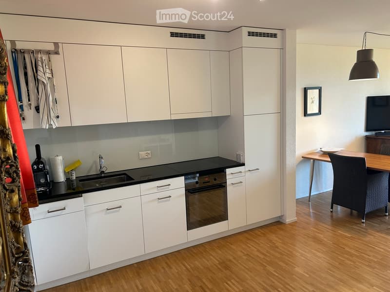 Modern kitchen with white cabinets, black countertops, and a wooden floor. Includes a coffee maker, oven, sink, and a small dining area with a table and chair.