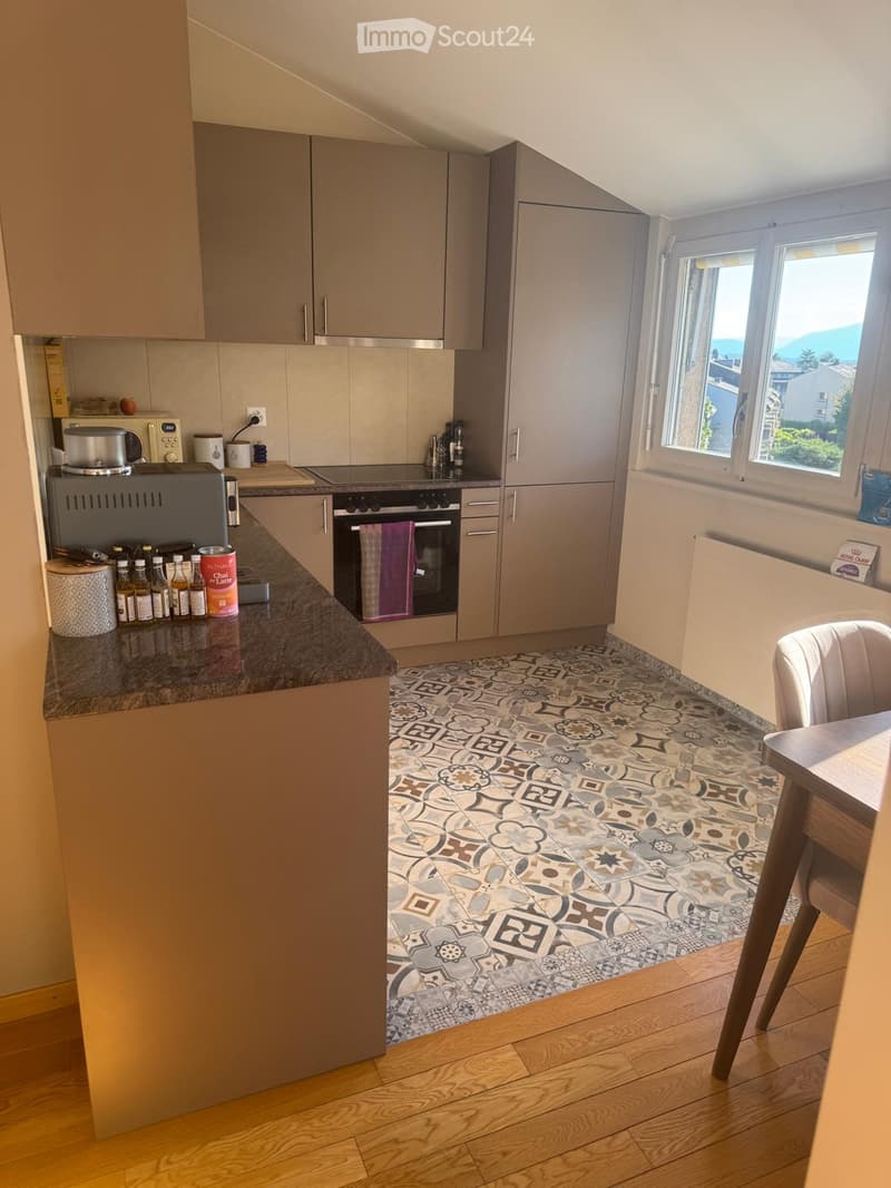 Modern kitchen with grey cabinets, granite countertop, and a patterned tile floor. Includes an oven, microwave, and a small dining area with a table and chair.