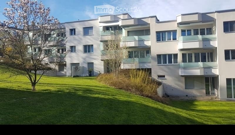 Modern multi-story residential building with white facade, multiple balconies, and surrounding green lawn.