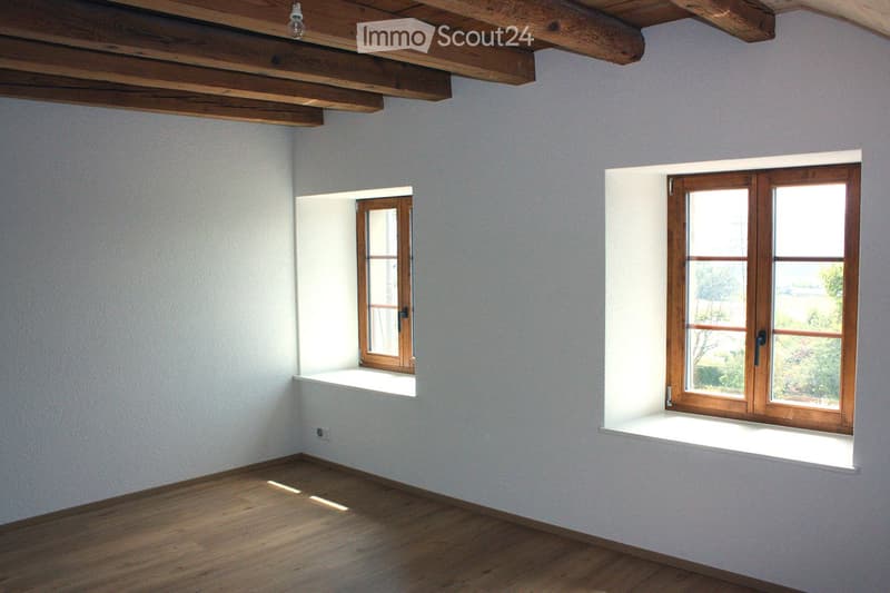 Empty room with wooden flooring, white walls, and two windows with wooden frames. The room features exposed wooden beams on the ceiling and a small built-in shelf beneath one window.