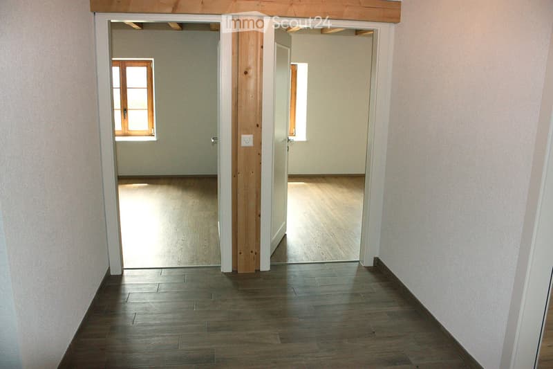 Empty hallway with wooden floors, white walls, and two open doors leading to adjacent rooms. Natural light streams in from windows in the adjacent rooms.
