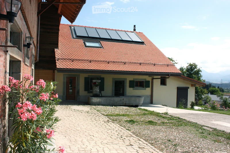 Two-story house with red-tiled roof, solar panels, and a small garden area with pink flowers. The house has a light yellow exterior with dark shutters and a stone pathway leading to the entrance.
