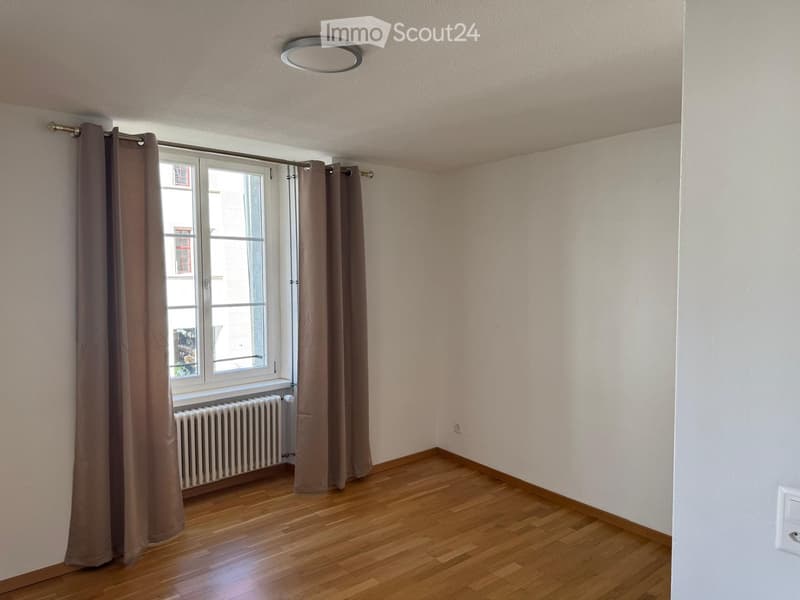 Empty room with wooden flooring, white walls, large window with brown curtains, radiator, and a ceiling light fixture.