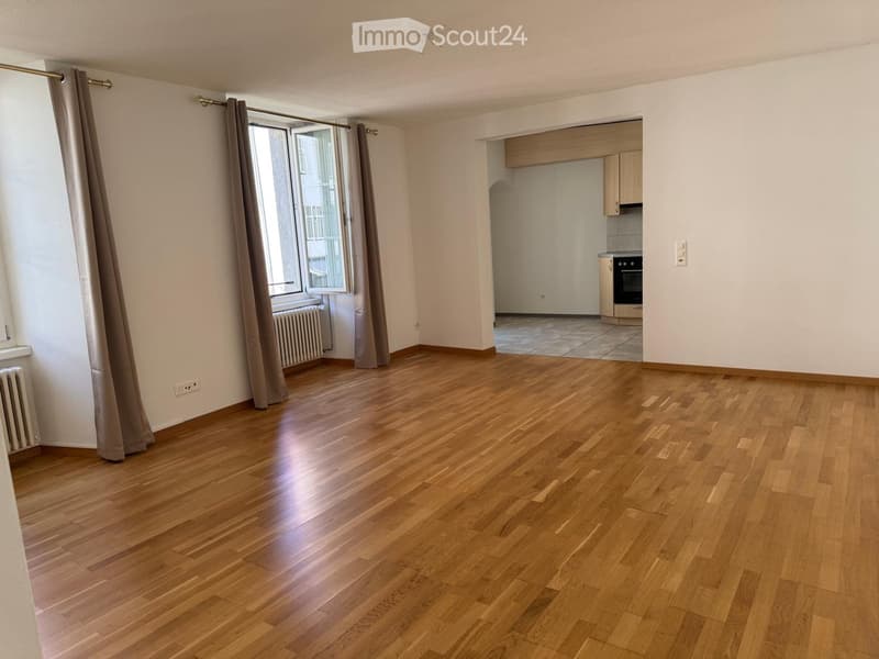 Empty room with wooden flooring, large window with curtains, radiator, and an adjacent kitchen area with cabinets and appliances.