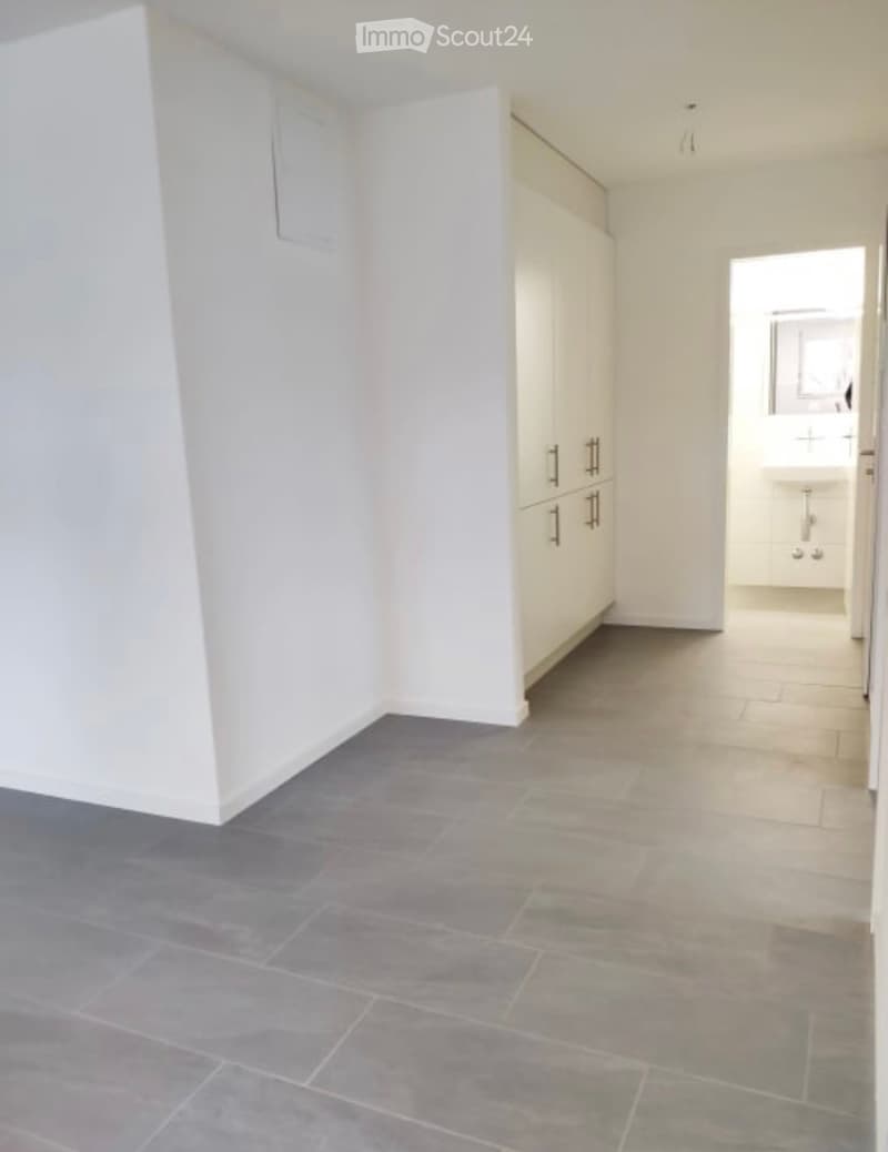 Empty room with grey tiled floor, white walls, and built-in cabinets. A doorway leads to a small bathroom area with a sink.