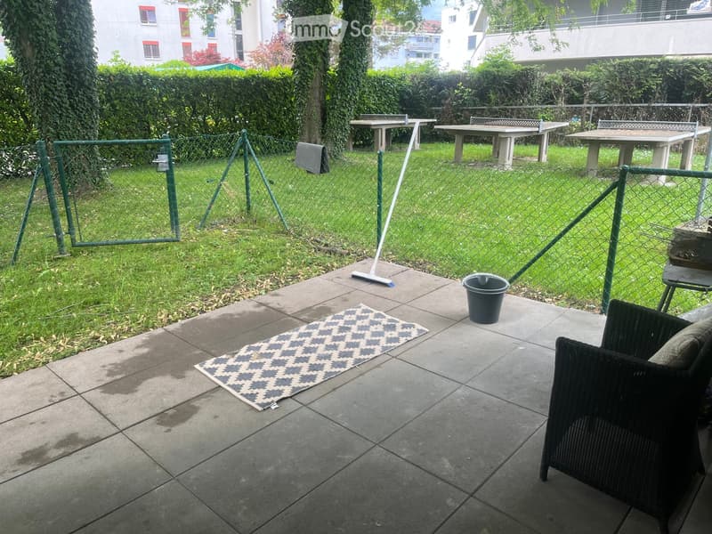 Outdoor patio area with tiled floor, a black wicker chair, a small rug, a broom, and a bucket. A fenced yard with tables and greenery is visible in the background.