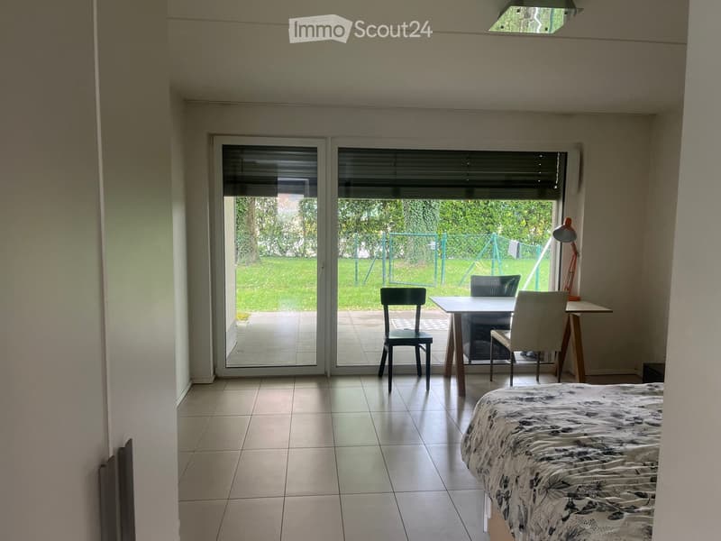Bright bedroom with tiled floor, large sliding glass doors leading to a patio, bed with patterned bedding, small table with chairs, and a desk with a lamp.
