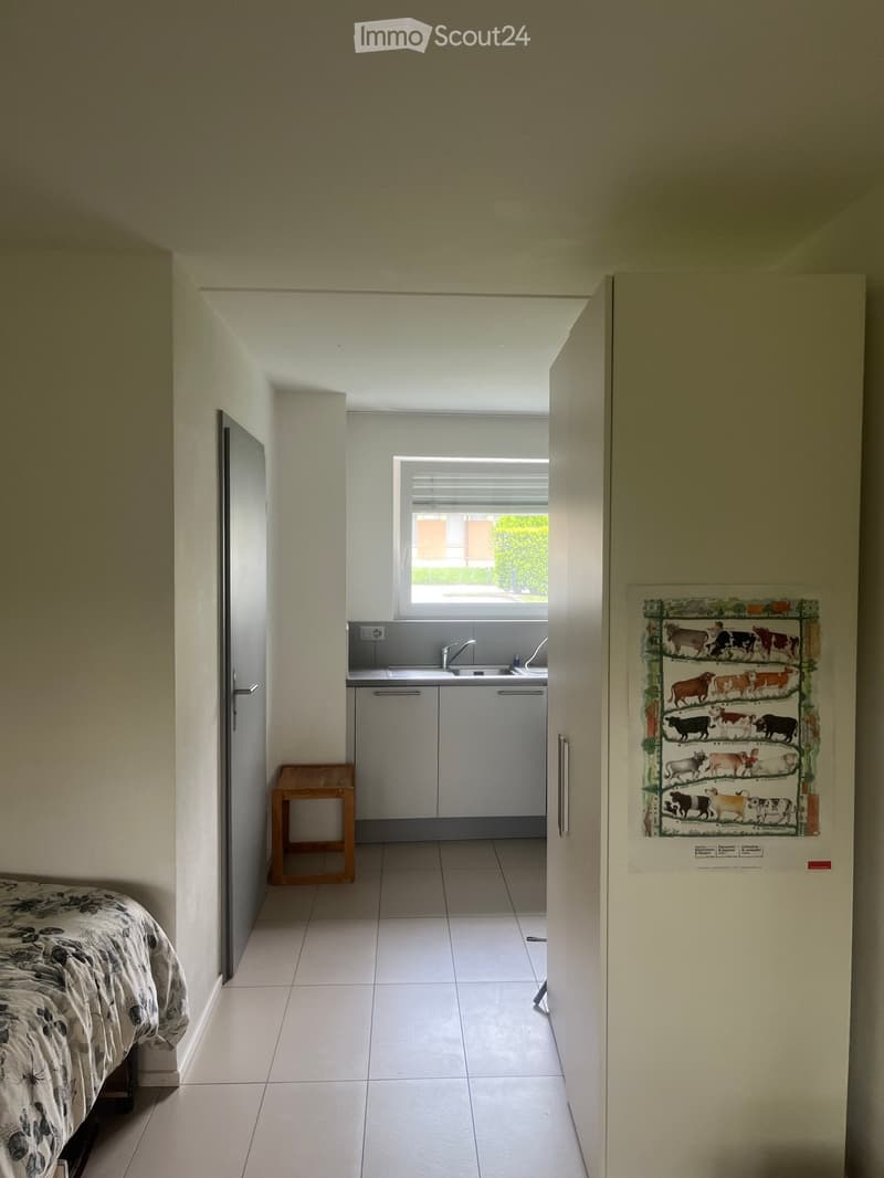 A furnished room with a bed, a small wooden stool, a refrigerator with a decorative poster, and a small kitchen area with a sink and faucet.