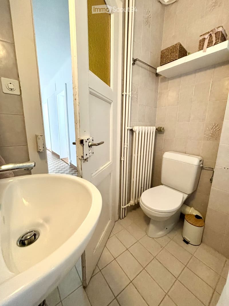 A small bathroom with a bathtub, toilet, radiator, and a trash bin. The walls are tiled, and there is a shelf with decorative items visible in the background.