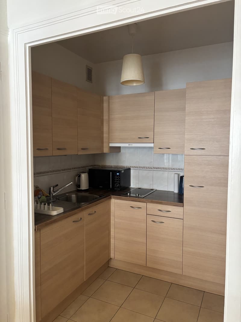 Modern kitchen with light wood cabinets, stainless steel sink, microwave, electric kettle, and induction stove. Beige tiled floor and walls.