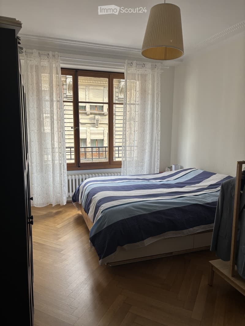 Cozy bedroom with wooden flooring, large window with curtains, bed with blue and white bedding, radiator, and wardrobe.