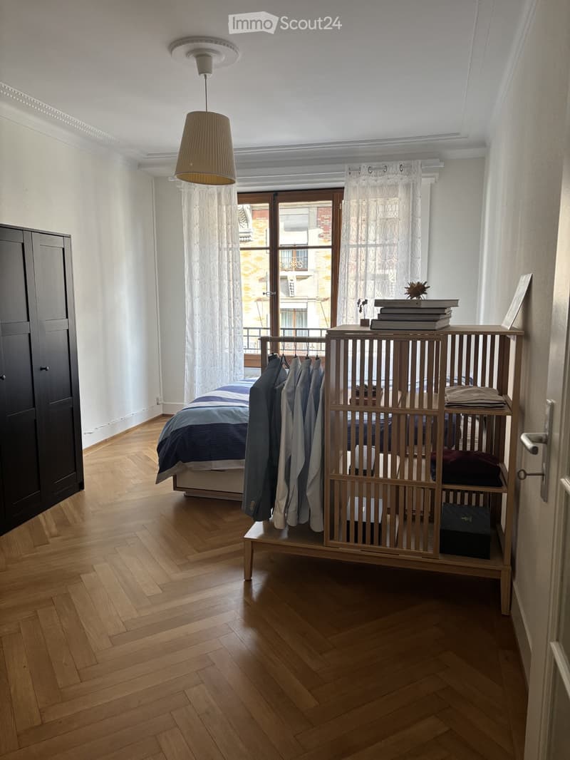 Modern bedroom with wooden herringbone flooring, a bed with blue and white bedding, a wardrobe, a hanging rack with clothes, and a curtained window with a view of a balcony.